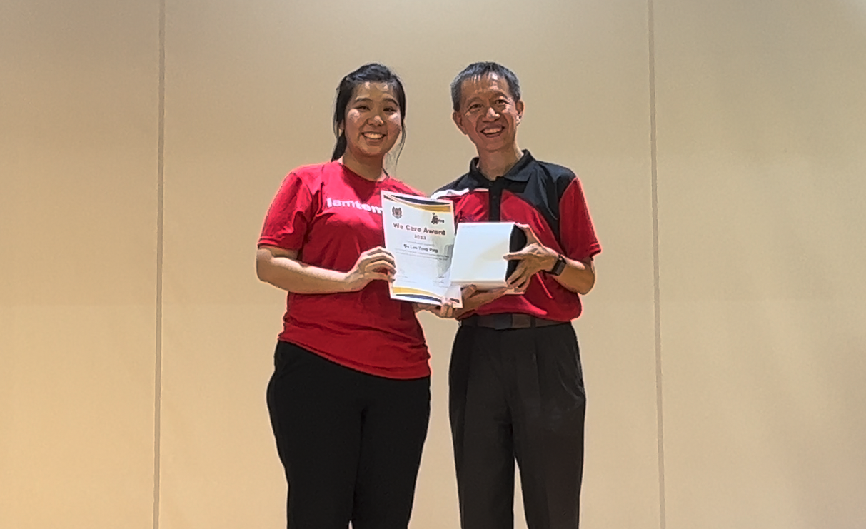 Two smiling people in red shirts hold a We Care Award certificate together in front of a beige wall.